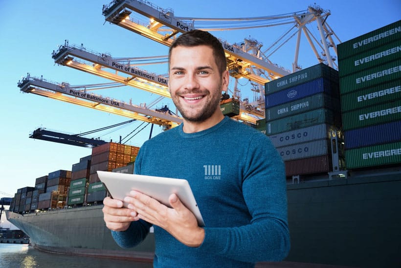 Mitarbeiter mit Tablet steht lächelnd vor einem mit Seecontainern beladenen Schiff